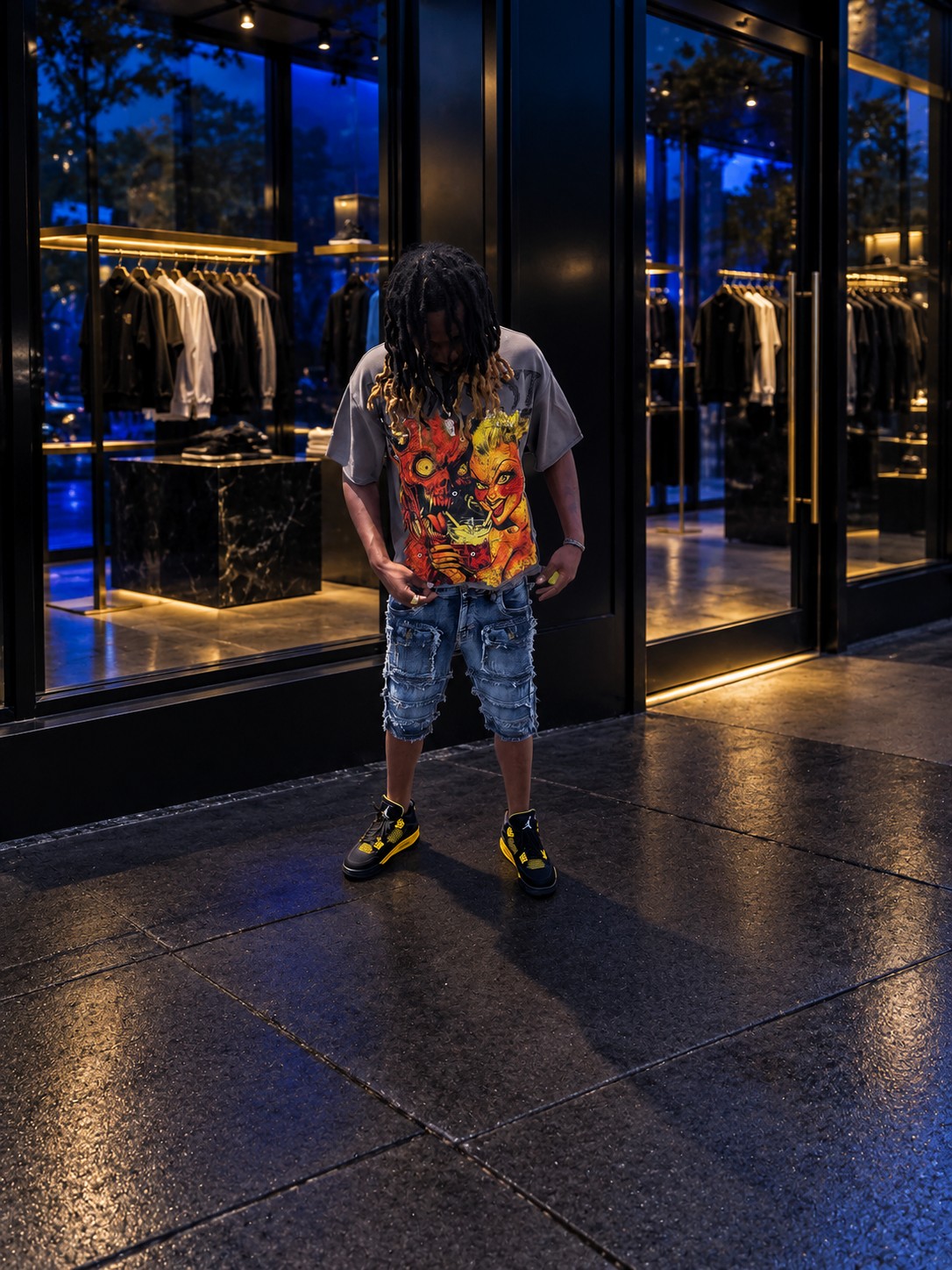 Streetwear look with a colorful graphic tee, distressed denim shorts, and black and yellow sneakers in front of a blue-lit clothing boutique